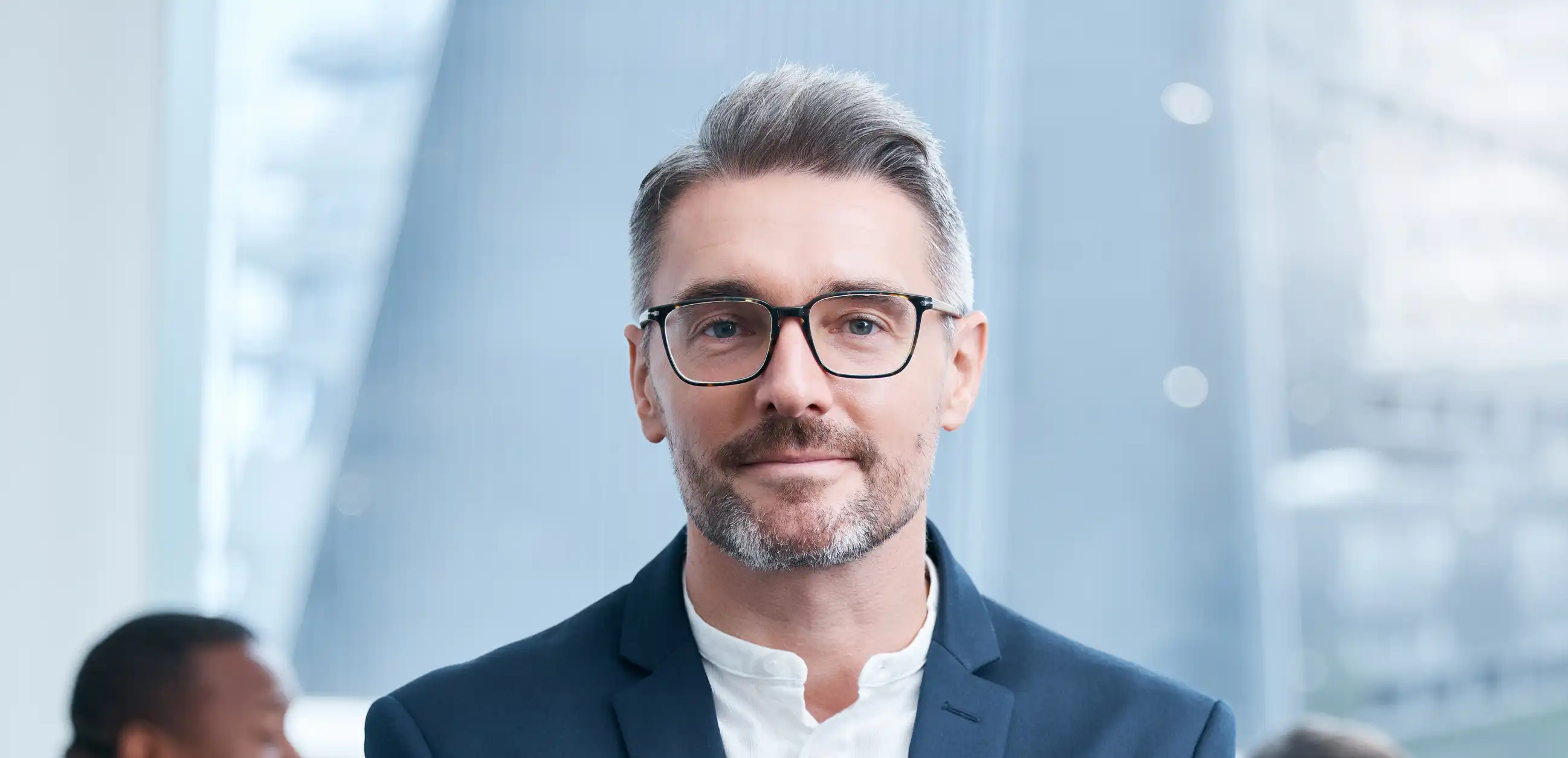 professional businessman wearing glasses folding arms looking at camera with two work colleagues behind him