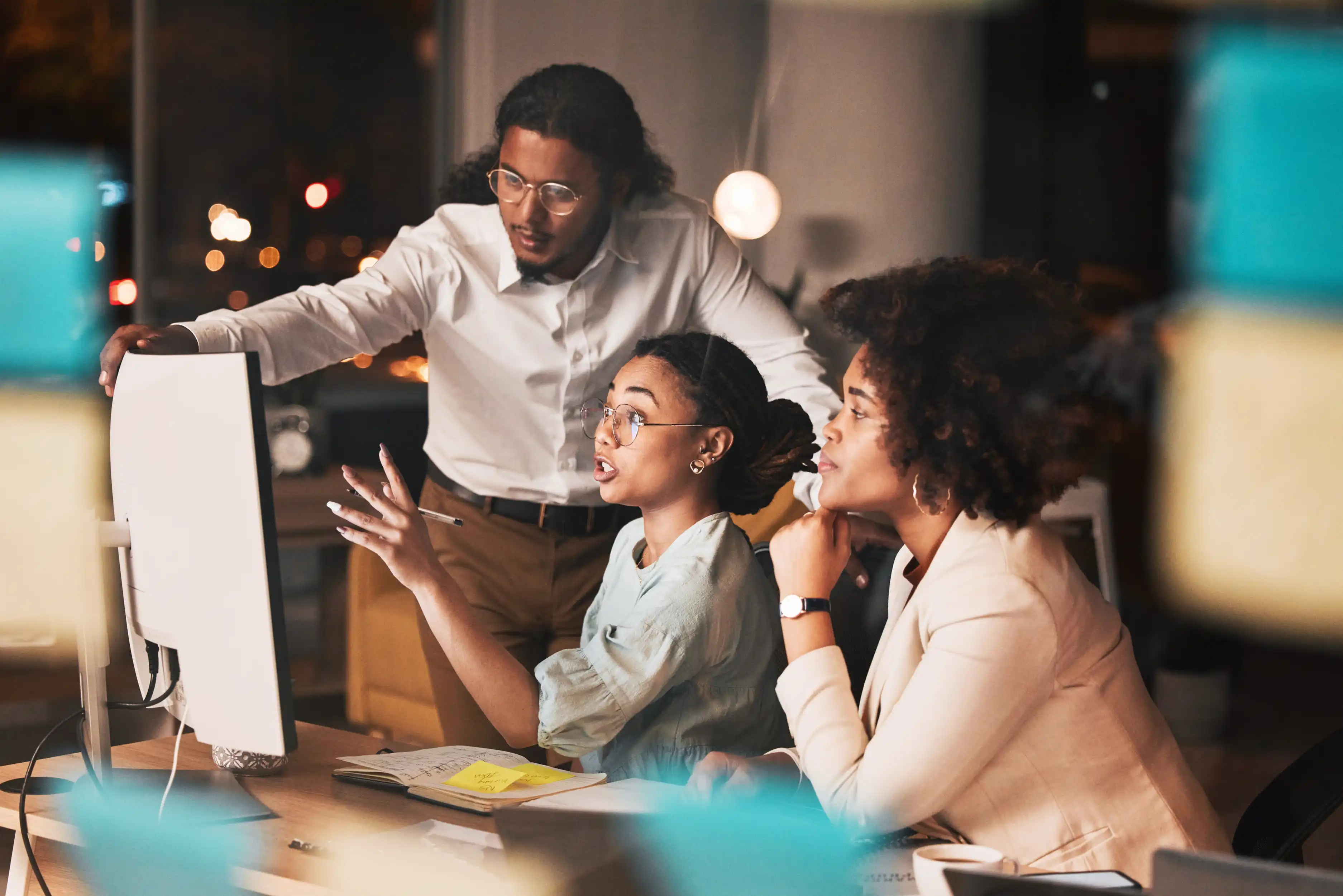 Three work colleagues conversing with each other in dimly lit office while viewing computer screen