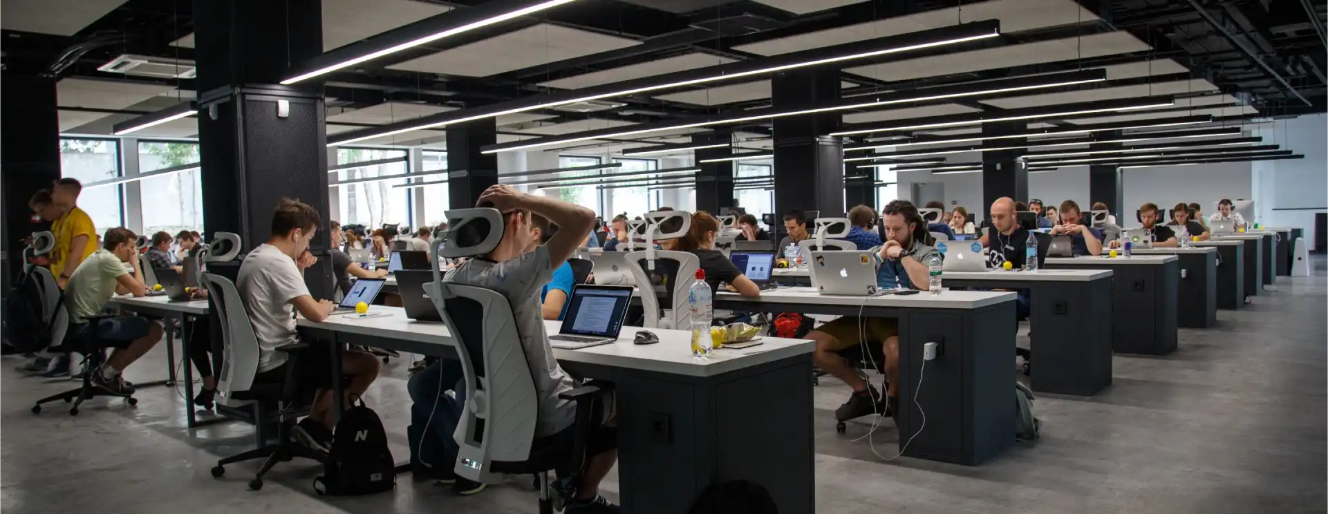 Several workers at apple computers sat on long row tables in a large room with LED lighting overhead