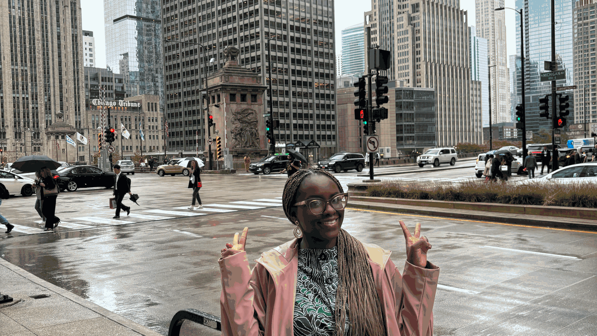 Standing at a rainy downtown Chicago intersection near the Chicago Tribune tower, wearing a pink raincoat with skyscrapers in the background