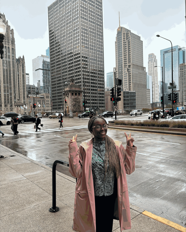 Standing at a rainy downtown Chicago intersection near the Chicago Tribune tower, wearing a pink raincoat with skyscrapers in the background