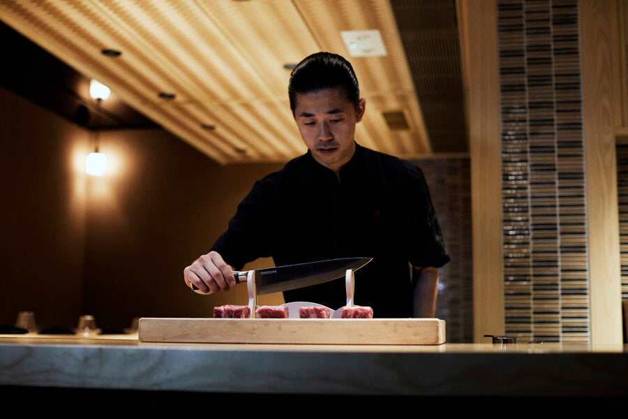 A Japanese chef demonstrates precise knife skills using a specialized cutting board with vertical supports for meat.