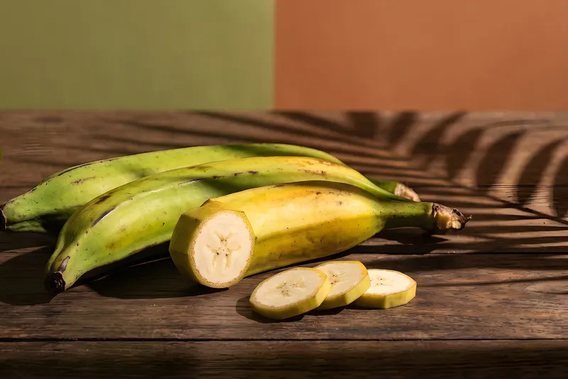 Long angular Nendran plantains on weathered teak, one cut open to show the pale flesh, with raw slices and a banana leaf
