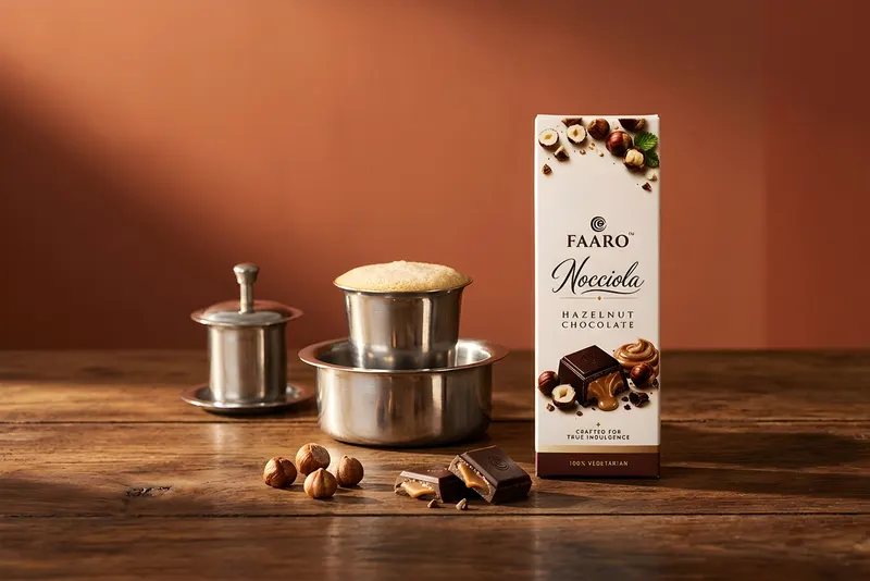A Faaro Nocciola hazelnut chocolate bar on teak beside a South Indian steel filter coffee davara and tumbler