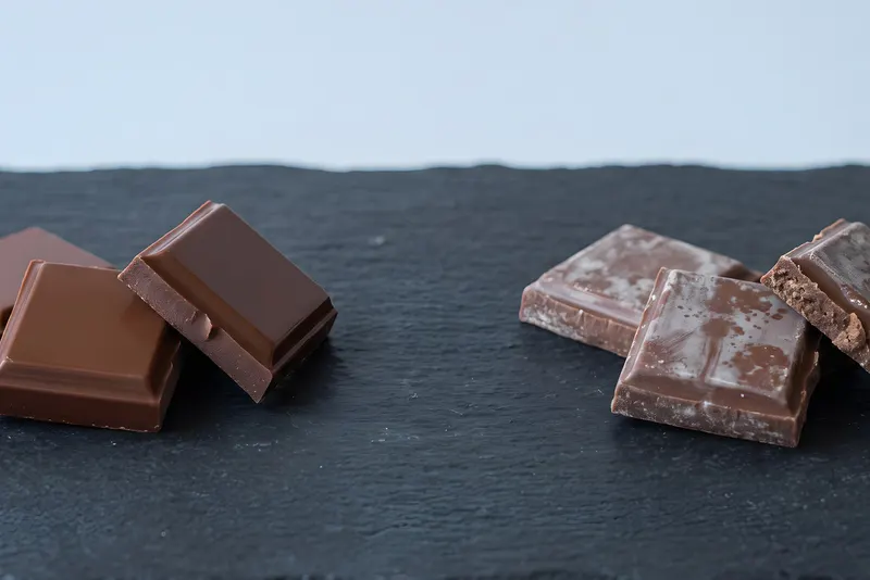 Glossy milk chocolate squares on the left beside dull, grey-streaked bloomed chocolate squares on the right, on a dark slate surface