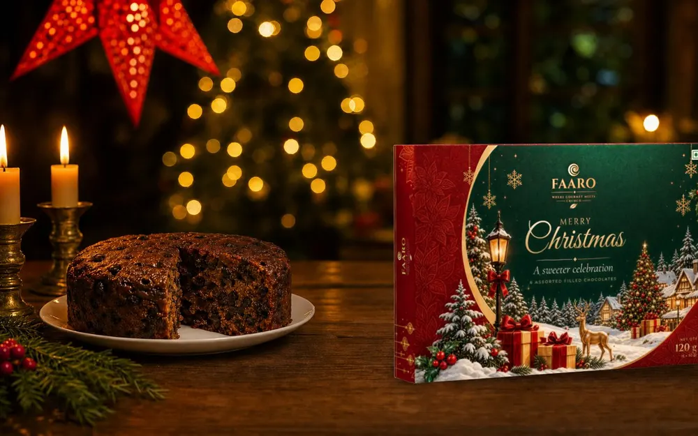The green, red and gold FAARO Merry Christmas gift box standing on a walnut table beside a cut plum cake and lit candles in brass holders, with a red paper star lantern and fairy-light bokeh behind