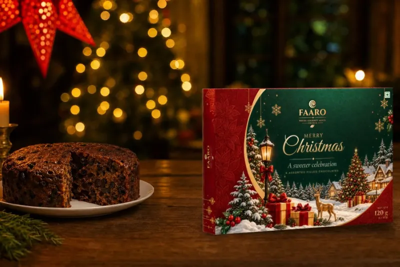 The green, red and gold FAARO Merry Christmas gift box standing on a walnut table beside a cut plum cake and lit candles in brass holders, with a red paper star lantern and fairy-light bokeh behind