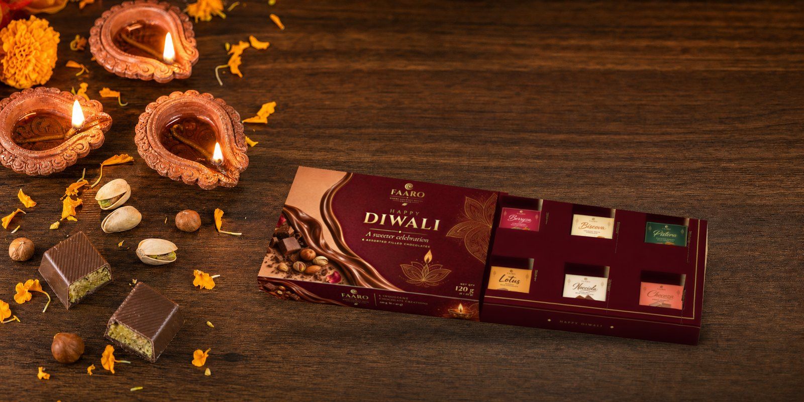 The FAARO Diwali gift box with its drawer open, showing six stickered filled chocolates, on a walnut table with lit terracotta diyas, marigold petals, pistachios and halved pistachio chocolates