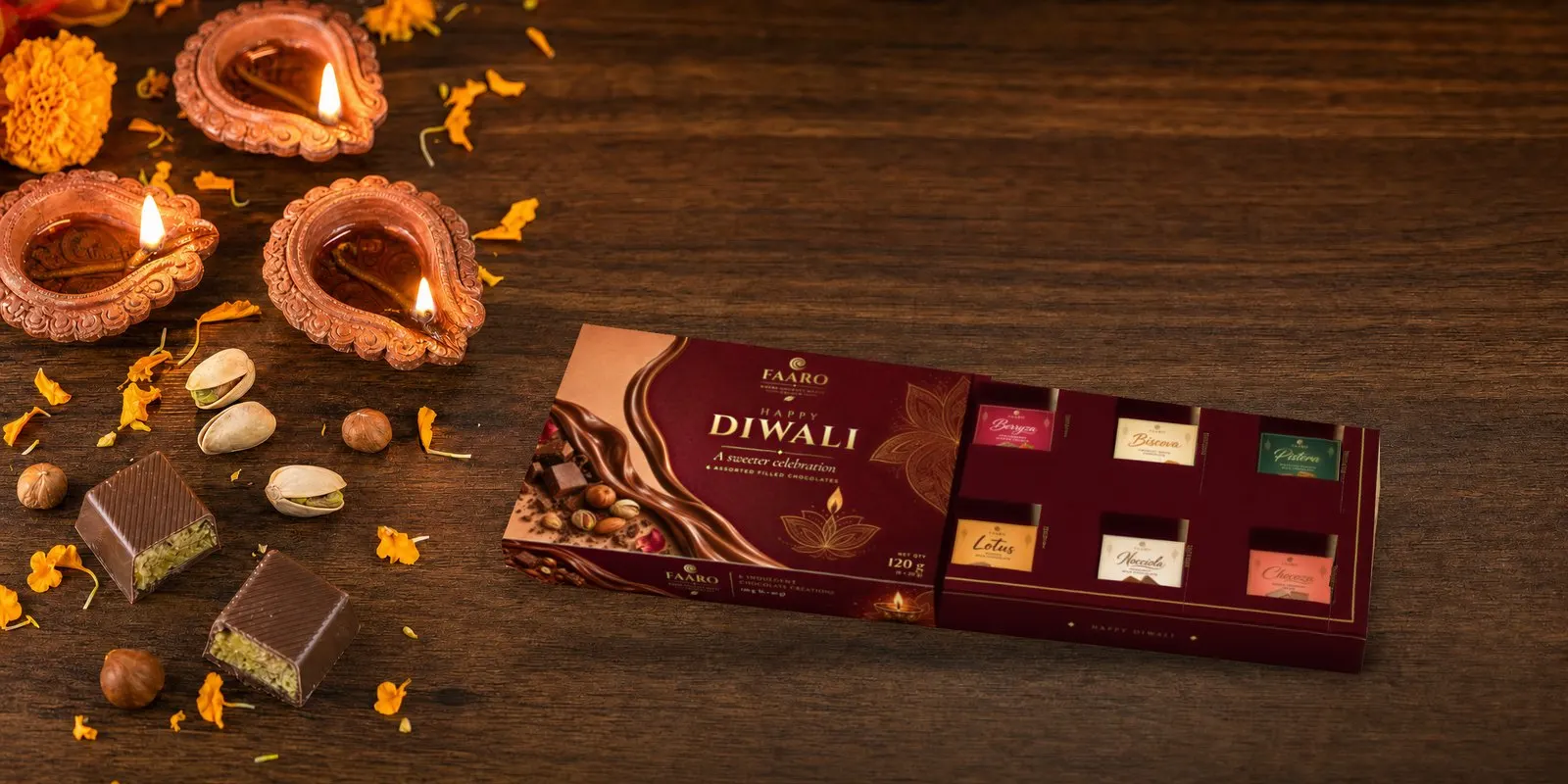 The FAARO Diwali gift box with its drawer open, showing six stickered filled chocolates, on a walnut table with lit terracotta diyas, marigold petals, pistachios and halved pistachio chocolates