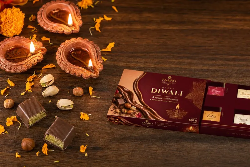 The FAARO Diwali gift box with its drawer open, showing six stickered filled chocolates, on a walnut table with lit terracotta diyas, marigold petals, pistachios and halved pistachio chocolates