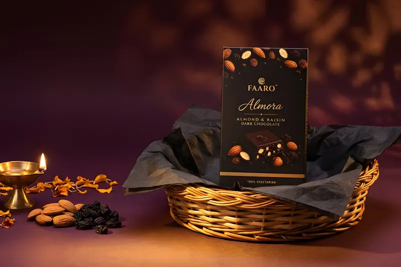 A Faaro Almora dark chocolate pack in an open woven Diwali hamper with a lit brass diya, almonds and marigold petals