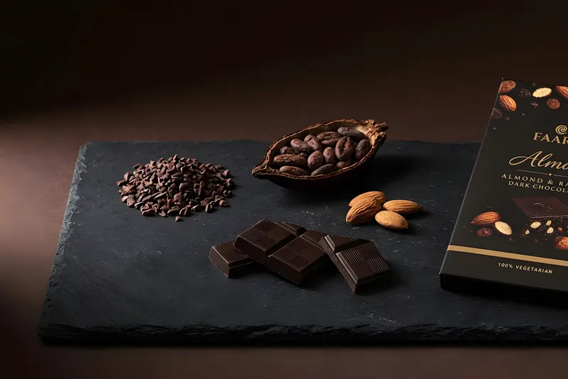 A Faaro Almora dark chocolate pack lying on dark slate with raw cocoa nibs, cocoa beans and almonds
