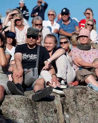 Publikum sitter tett i tett på svaberget og nyter konserten på Stangholmen under en strålende blå himmel.