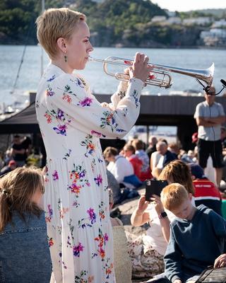 Trompetist og kunstnerisk leder Tine Thing Helseth spiller trompet foran et solfylt Stangholmen med publikum under blå himmel