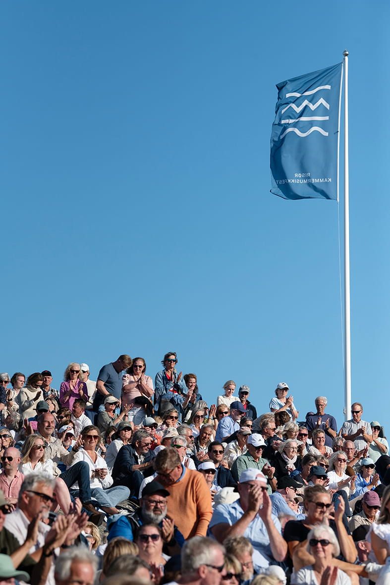 Risør kammermusikkfest sitt flagg vaier mot en blå himmel med publikum sittende i solskinnet