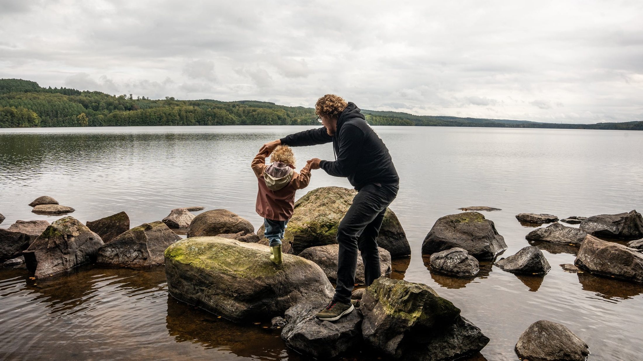 Joe leading his young daughter across boulders at the edge of a lake