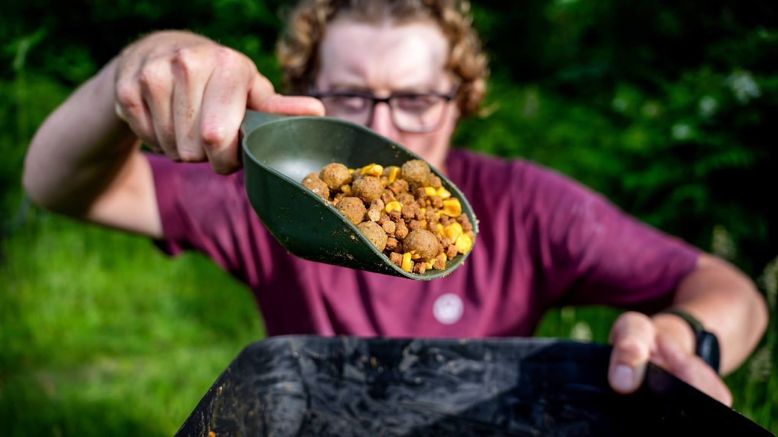 Joe holding out a scoop of boilies and sweetcorn over a bait bucket