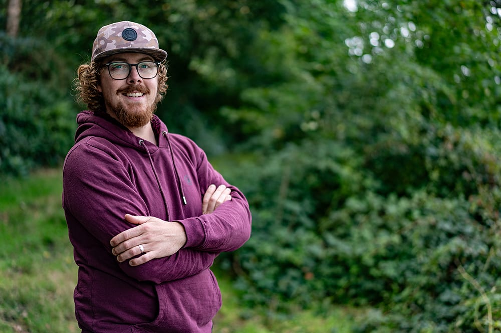 Joe Wooltorton, arms folded, smiling in a camo cap and burgundy hoodie in front of green woodland