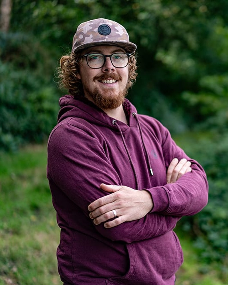 Joe Wooltorton, arms folded, smiling in a camo cap and burgundy hoodie in front of green woodland