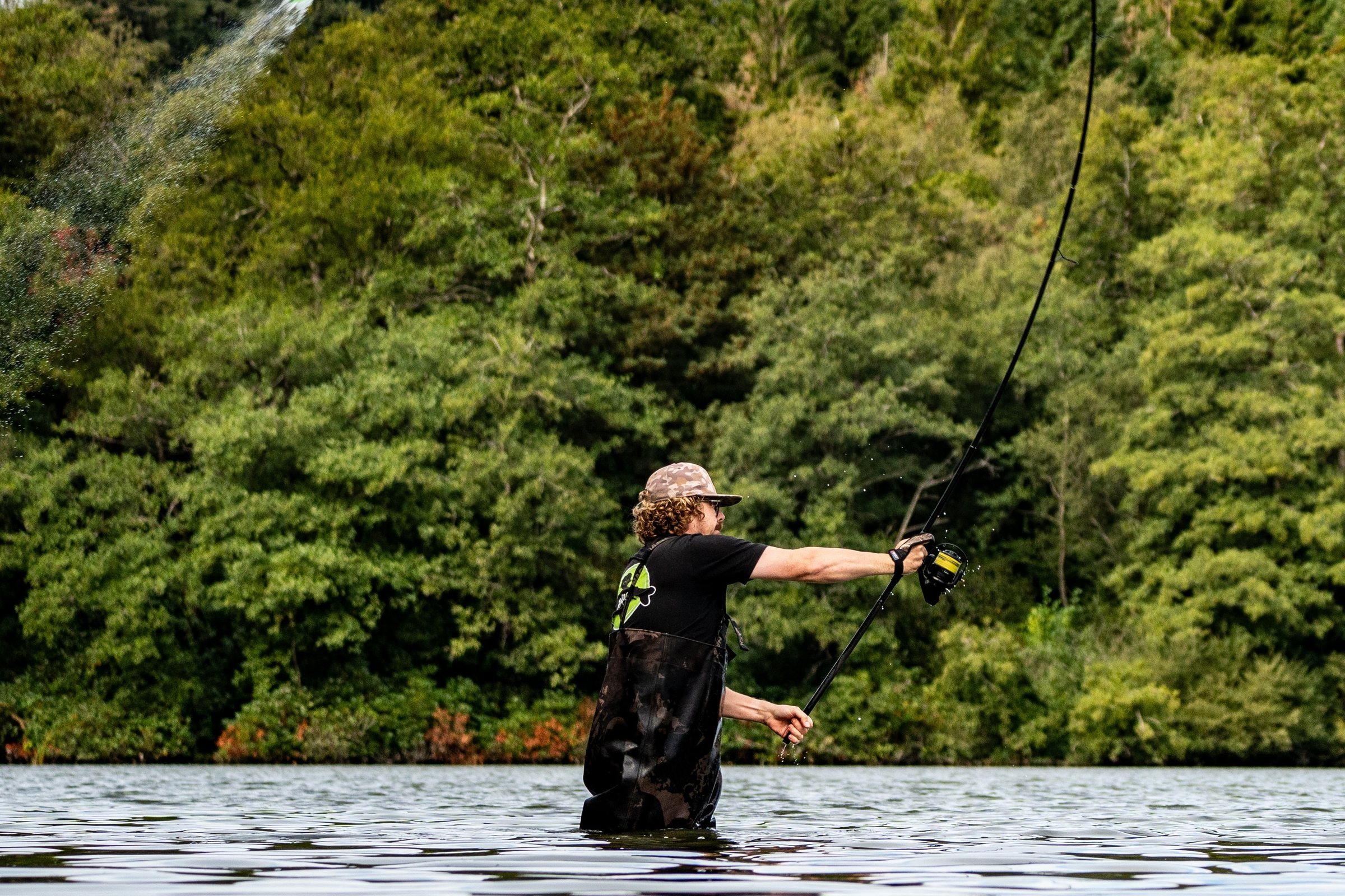 Joe mid-cast, wading in a lake, the rod bent against a wall of trees