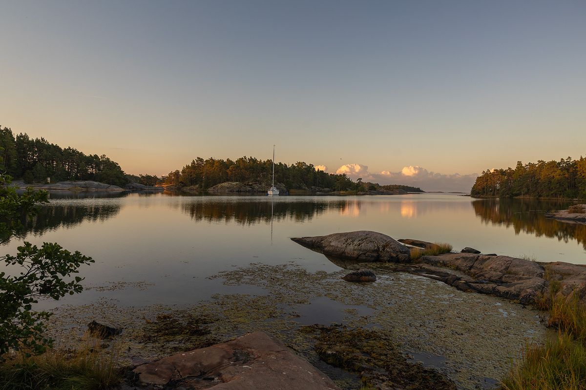 Stenar i förgrunden och segelbåt i spegelblank vik