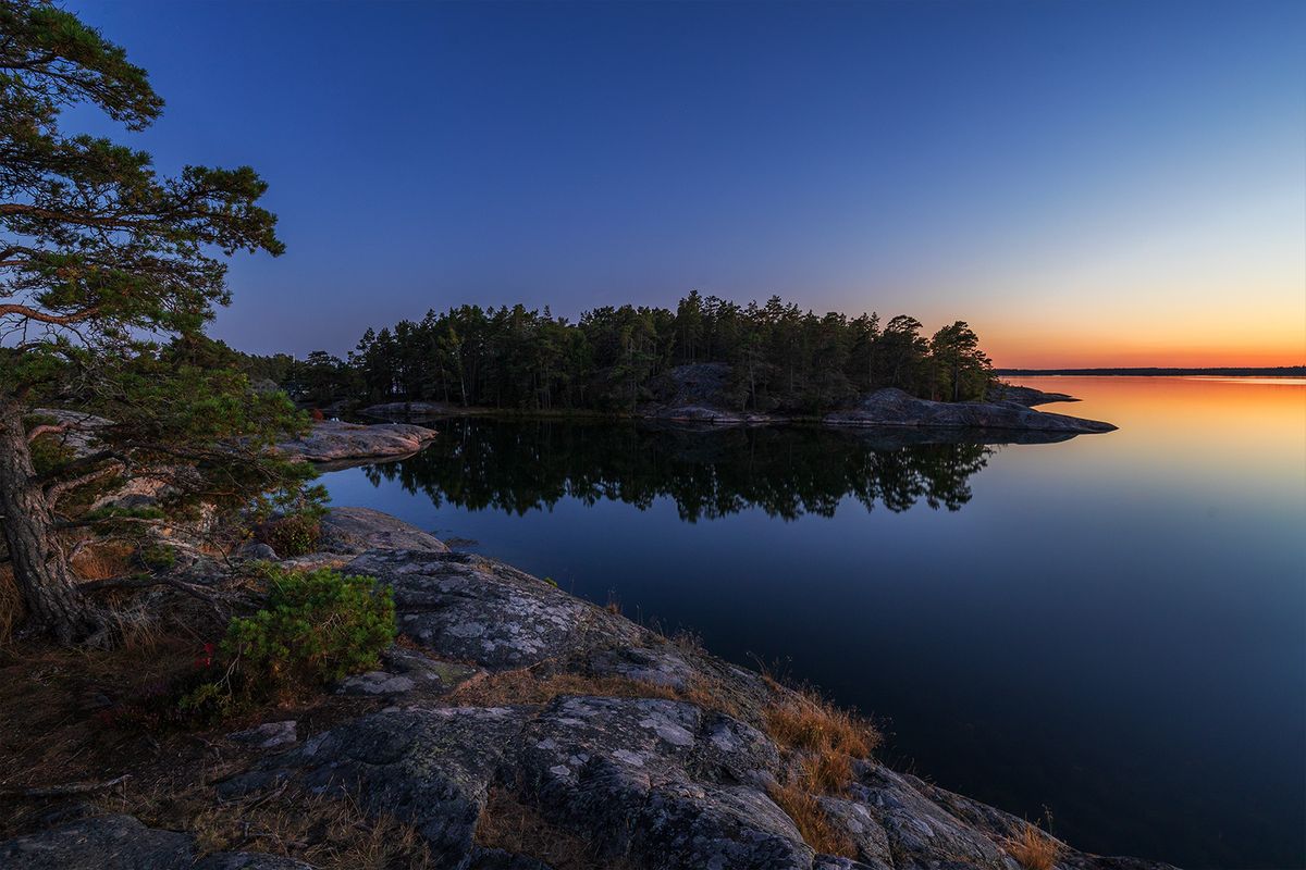 Liten skogö och spegelblankt vatten mot orange horisont i blå timmen