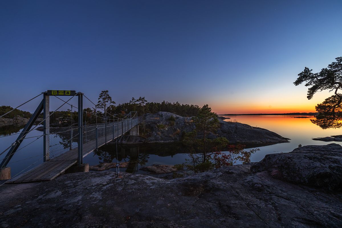 Hängbro över stilla vatten mot orange horisont i blå timmen