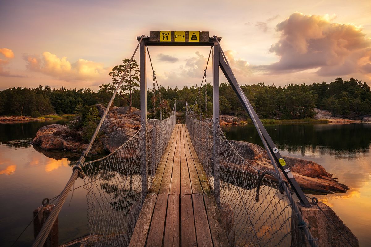 Träbro med vajrar leder mot en skogö i varmt kvällsljus