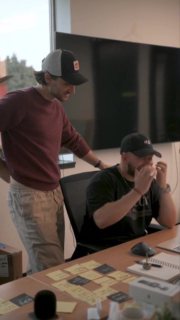 A man in a green shirt reaching towards a whiteboard mid-sentence, with three colleagues behind him listening, one holding a mug