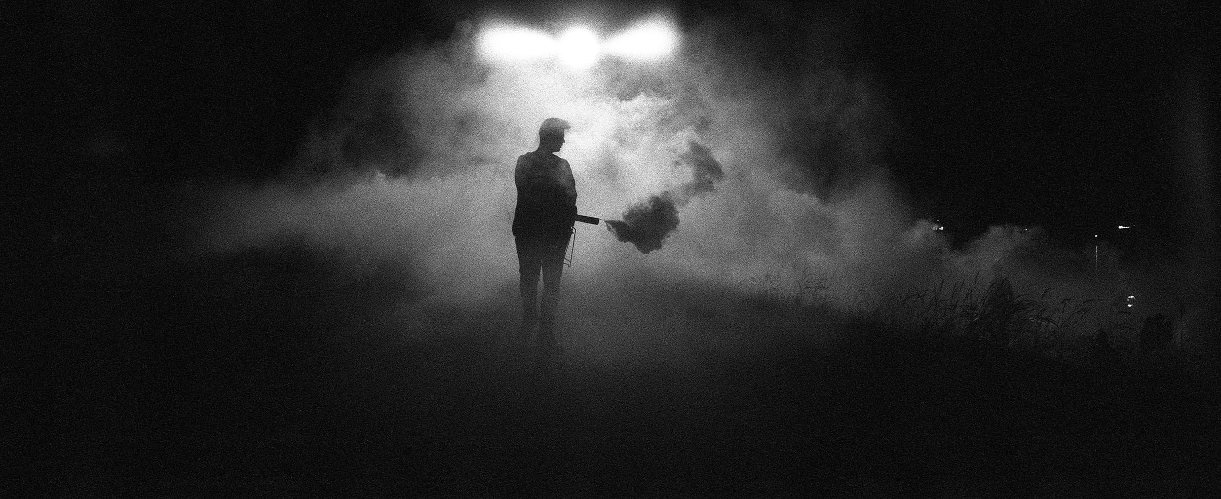 Een silhouet van een persoon die een rookfakkel vasthoudt, gevangen in een dramatische zwart-wit sfeer met dikke mist en fel licht.