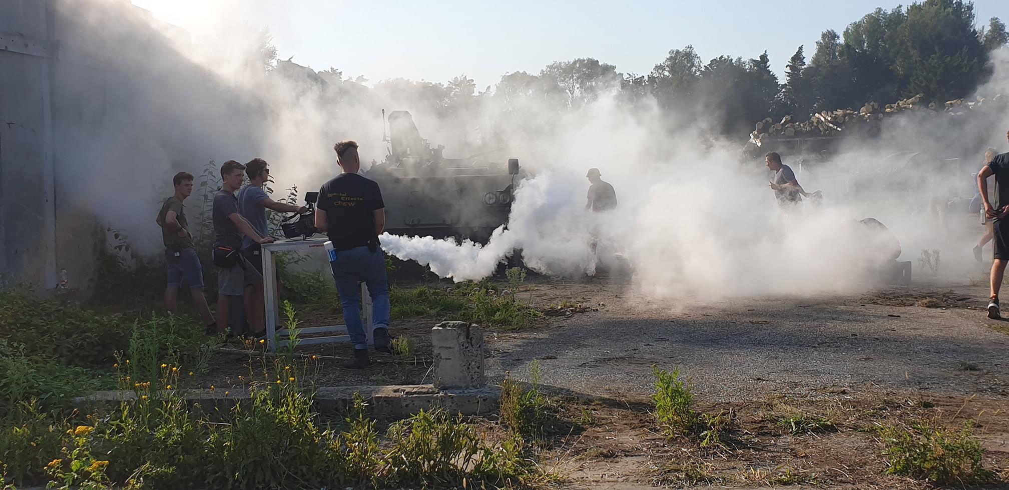 Een groep mensen werkt op een filmset waar rook wordt gegenereerd, terwijl een gepantserd voertuig in de achtergrond zichtbaar is.