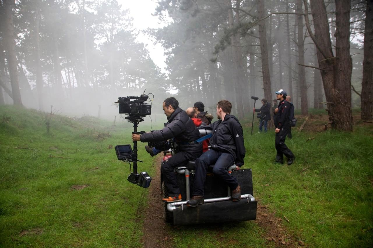 Een filmcrew rijdt op een camera-rig door een mistig bos, omringd door bomen, terwijl cameramannen de scène vastleggen.