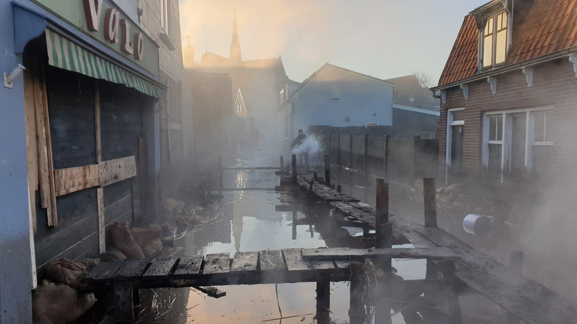 Een door mist en rook omhulde oude straat met vervallen houten bruggetjes en zandzakken, wat een sfeer van mysterie en verlatenheid creëert.