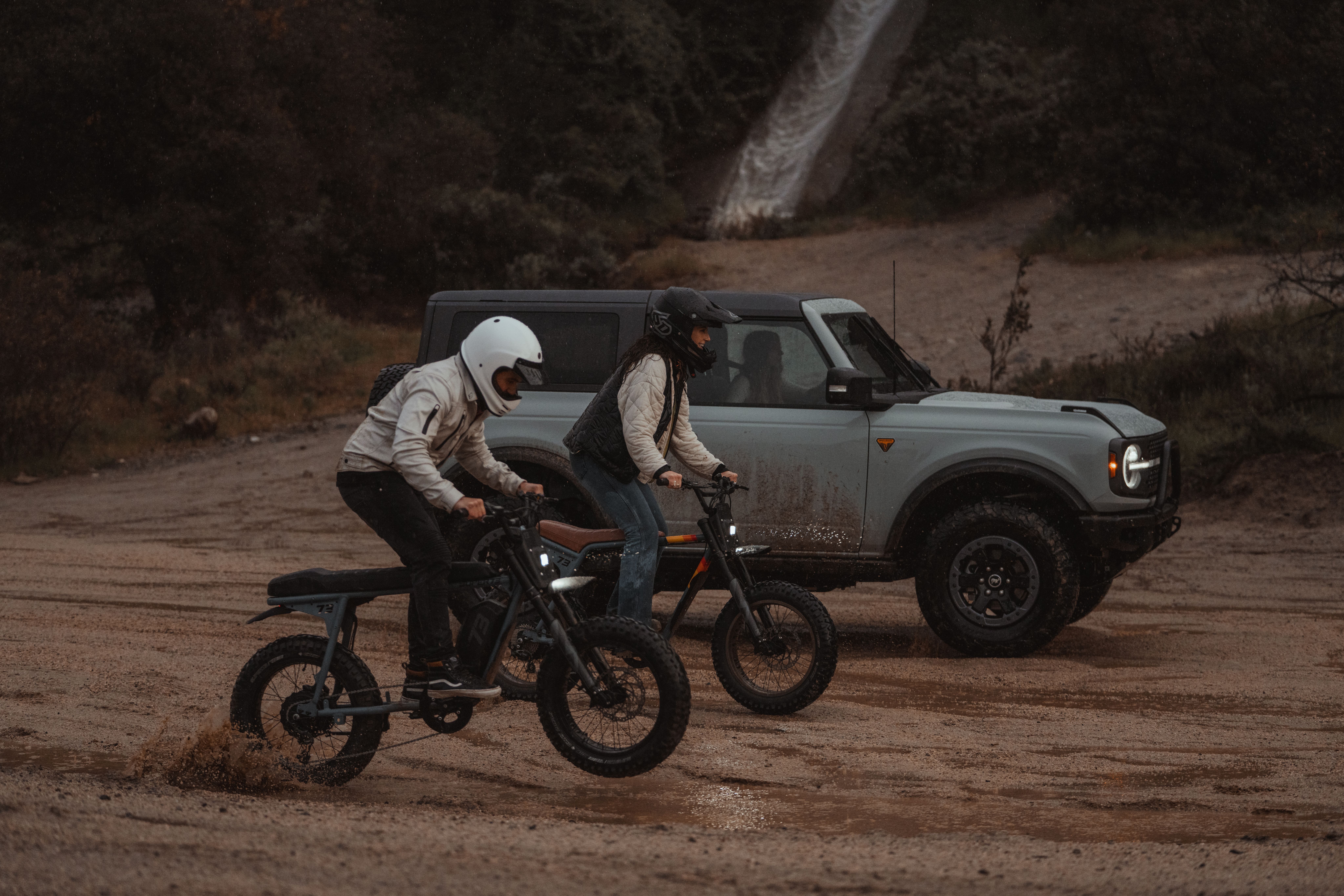 Two electric bikes riding in the mud next to a bronco truck