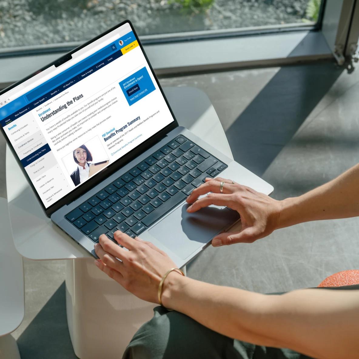 Overhead view of a person's hands typing on a laptop displaying an insurance benefits website with the heading 'Understanding the Plans' and 'Benefits Program Summary' visible on screen. The person is seated at a white round table near a window.