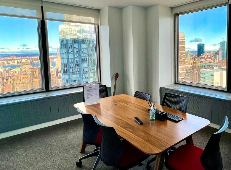 A bright conference room with large windows on two walls showing panoramic city skyline views. The room features a wooden oval conference table surrounded by black mesh office chairs, with a guitar visible in the corner. The space has white walls and demonstrates a modern office environment with natural lighting.