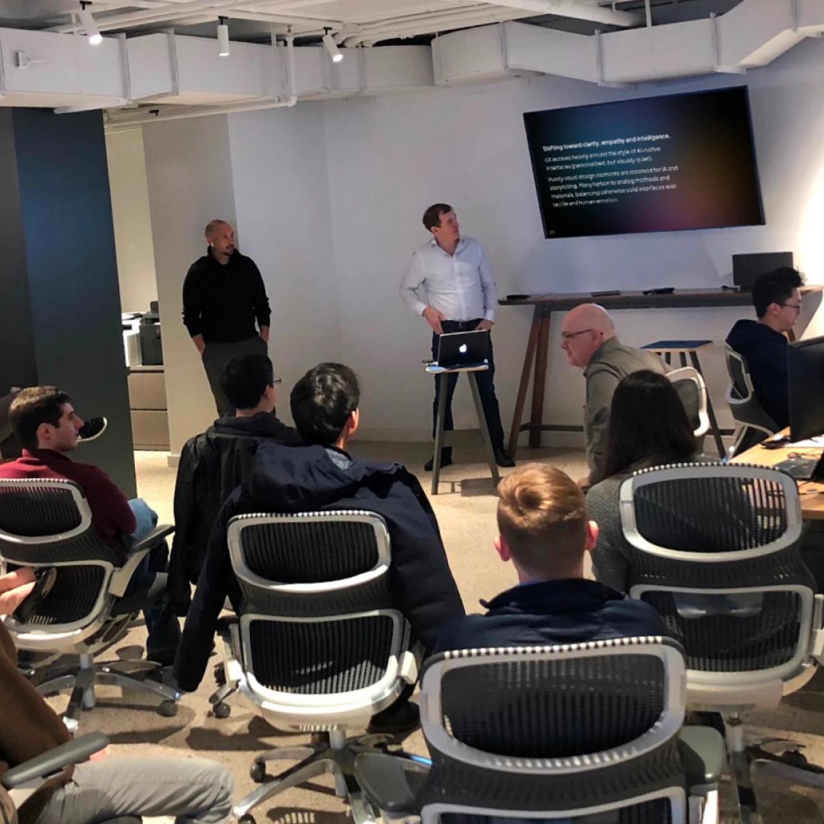 A business presentation taking place in a modern office space with white walls and exposed ceiling. Two presenters stand at the front - one in a black shirt and one in a white button-down with a laptop on a stand. An audience of approximately 10 people sits in ergonomic office chairs with black mesh backs and white frames. A large monitor displays text on the wall behind the presenters.