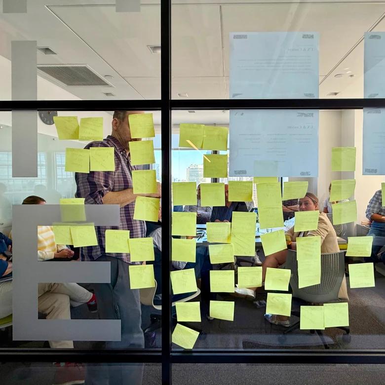 View through a glass wall into a modern office meeting room where team members collaborate around a table covered with numerous yellow sticky notes. A person in a plaid shirt stands at the glass, placing additional sticky notes, while others are seated working on laptops. The bright, minimalist space features white walls and ceiling-mounted ventilation.