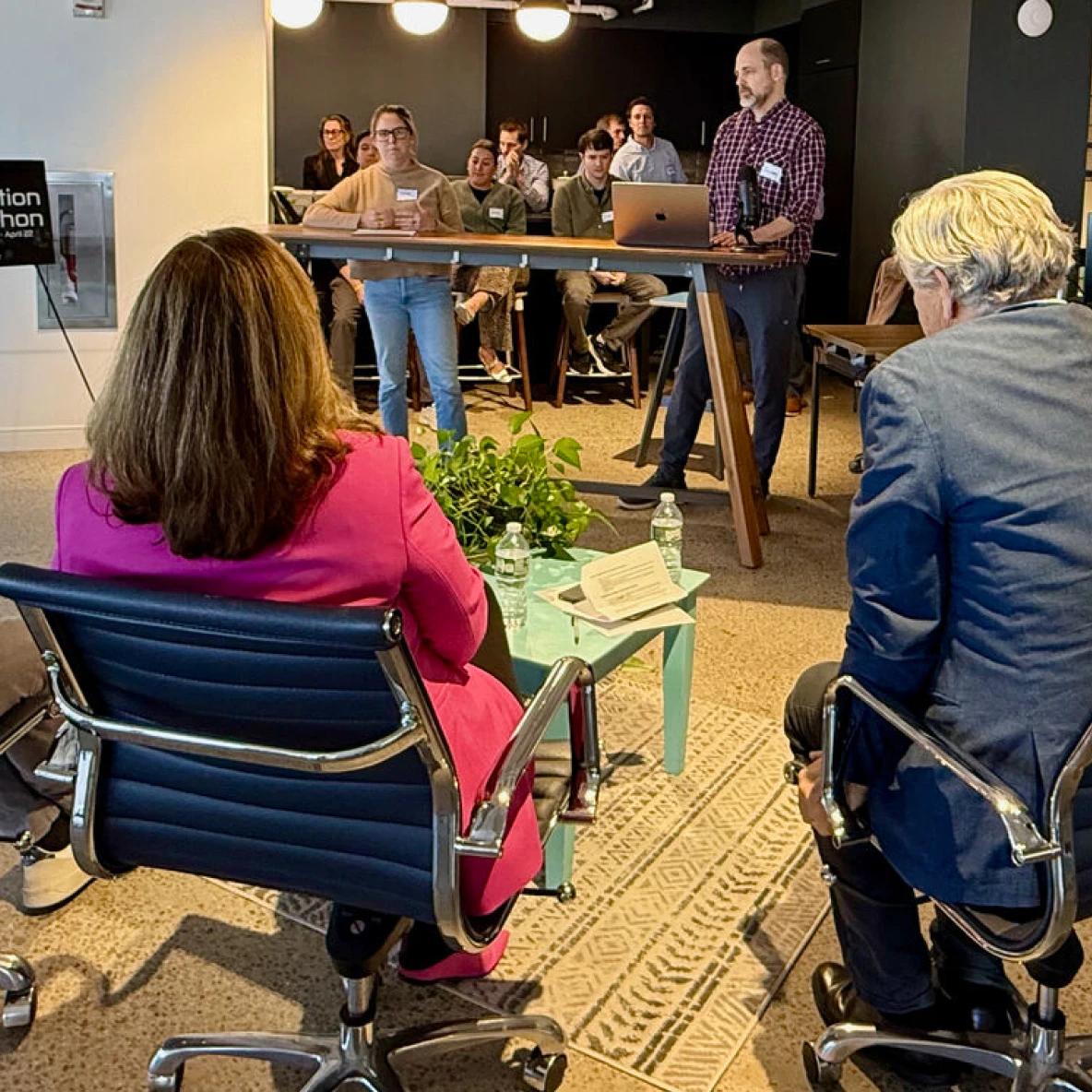 A community workshop or meeting in progress, with a presenter in a plaid shirt standing at a high table addressing attendees. In the foreground, two people are seated in office chairs, one wearing a bright pink blazer. Several participants are visible at the table in the background. The modern office space features pendant lighting and a patterned area rug.