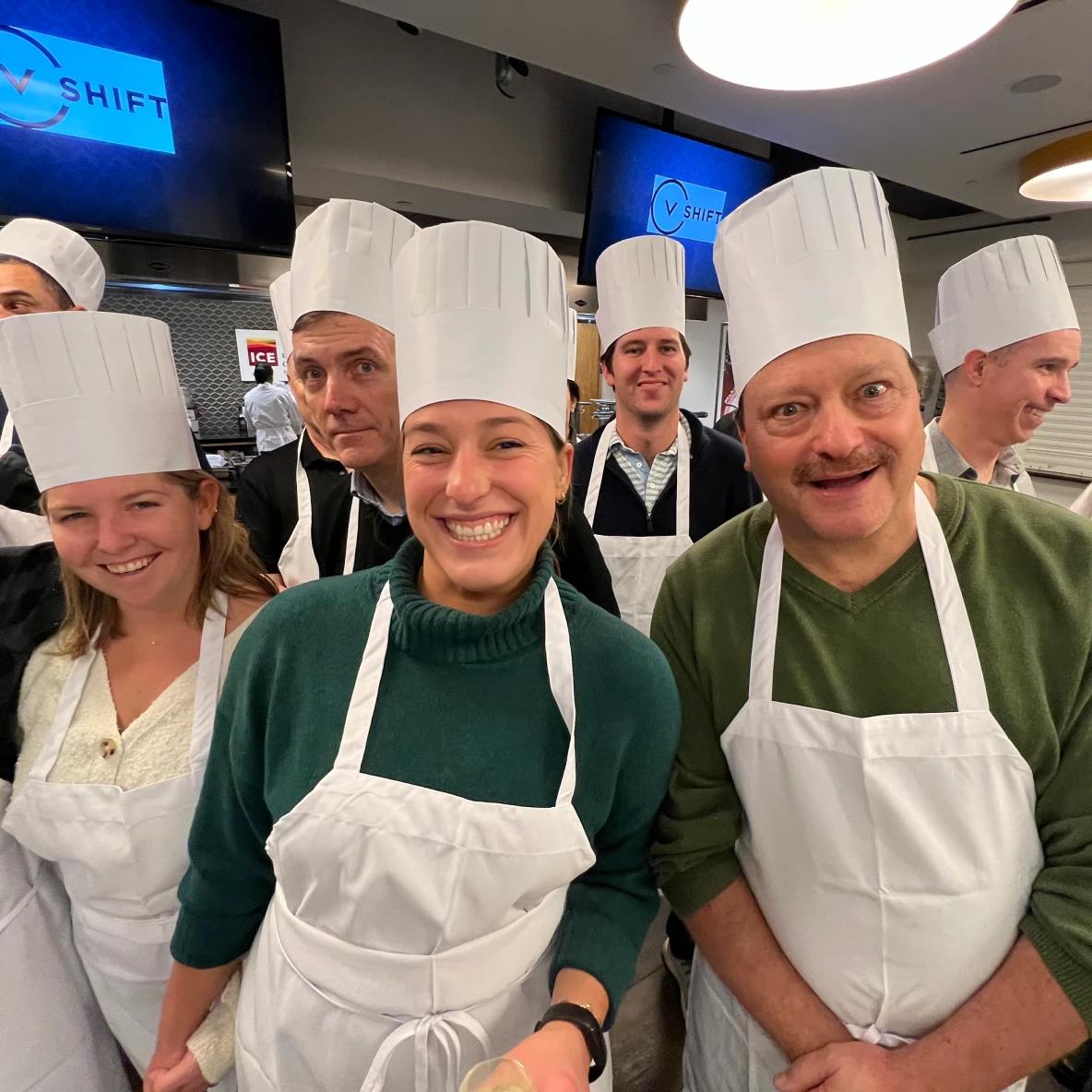 Group of seven people wearing white chef hats and aprons, smiling at the camera in a professional kitchen setting with SHIFT branding visible on screens in the background