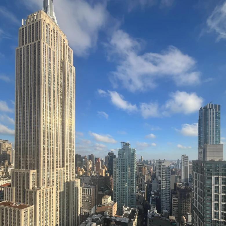 Daytime view of the iconic Empire State Building dominating the left side of the frame, with its distinctive Art Deco architecture and limestone facade. The Manhattan skyline extends to the right, featuring modern glass skyscrapers and mixed architectural styles under a bright blue sky with scattered white clouds.