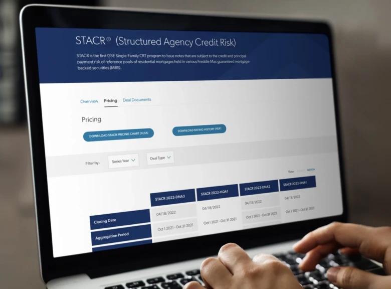 Close-up view of a laptop screen displaying the STACR (Structured Agency Credit Risk) pricing page. A hand is visible on the keyboard. The screen shows a data table with closing dates and aggregation periods for various STACR series from 2021-2023, along with download options for pricing charts and history at the top.