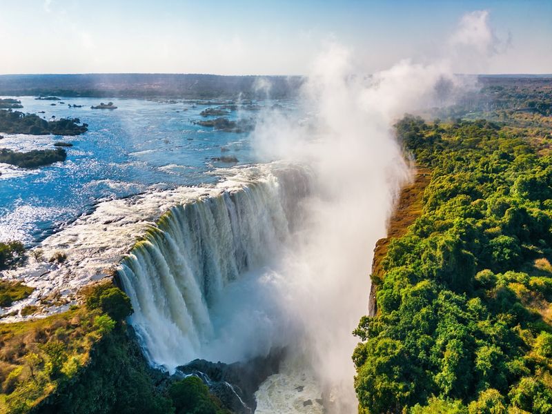 Mauritius and safari holiday, a colossal waterfall crashes over a rocky cliff, sending mist high into the air as the river winds through lush green landscapes under a bright blue sky.