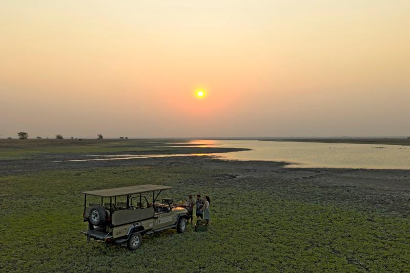 Guests pause beside a safari vehicle at sunset overlooking a winding waterway, capturing a moment of quiet connection shaped by the impact in Gorongosa.