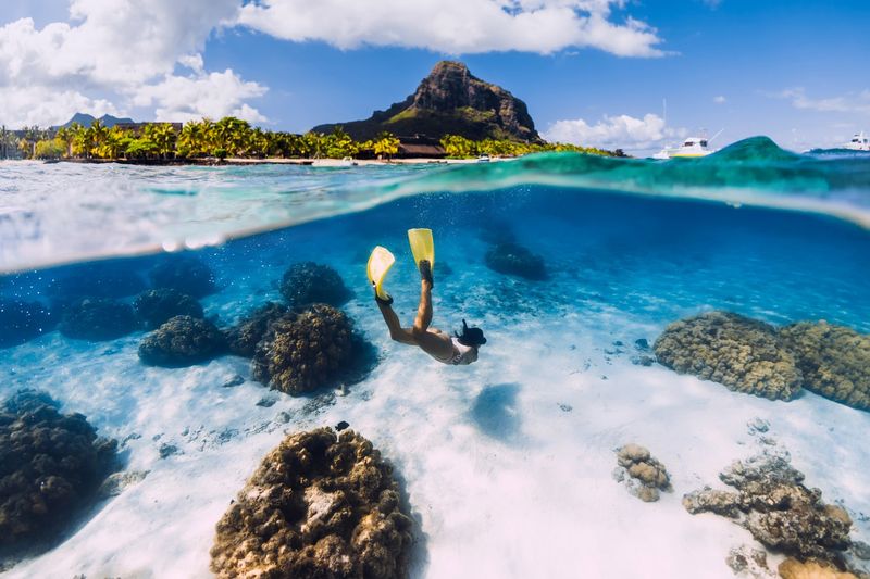 A snorkeler with bright yellow fins glides through crystal-clear turquoise waters above a vibrant coral reef, with Mauritius' lush coastline and towering Le Morne Mountain in the background.