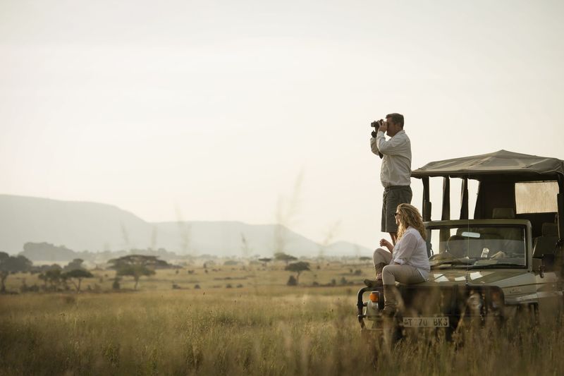 Guests on a Dunia Camp game drive vehicle in Tanzania’s Serengeti, with one person standing to photograph the open savannah while another sits on the bonnet overlooking the plains.