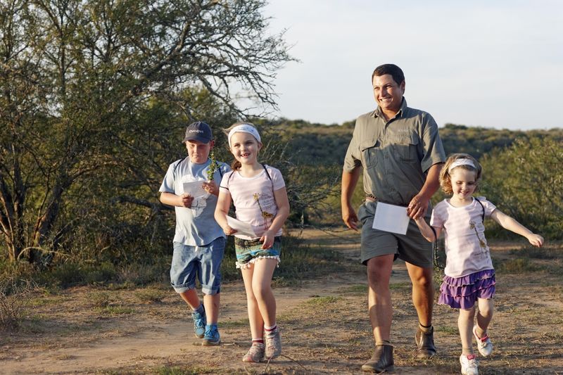 A smiling guide leads three children on a bush walk, with the kids eagerly holding activity sheets and looking engaged in their surroundings.