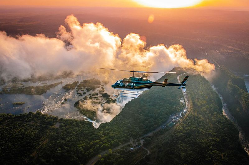 A helicopter hovers over golden mist and rainforest as it traces the edge of the best side of Victoria Falls at sunrise.