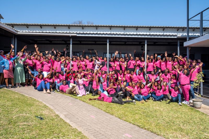 A massive group of GWF team members and students, dressed in bright pink, gather outside a building, raising their hands and cheering in celebration under the clear blue sky.