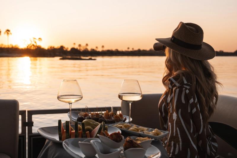 A woman in a wide-brimmed hat sips wine while watching the sunset over a tranquil river, with a table of gourmet bites and fresh fruit in front of her.